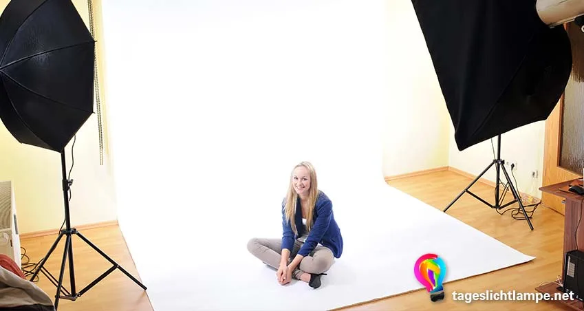 Einsatz einer Tageslcihtlampe im Fotostudio. Zu sehen sind links und rechts im Bild jeweils zwei Standlampen, die eine junge Frau ausleuchten, die auf einem weißen Hintergrund auf dem Boden sitzt.